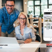 Why-Integrated-Security-Systems-Are-a-Smart-Investment Smiling professionals in an office setting collaborating on a laptop, with a smartphone displaying integrated security system features, emphasizing the importance of security solutions for businesses in Wilmington, DE.